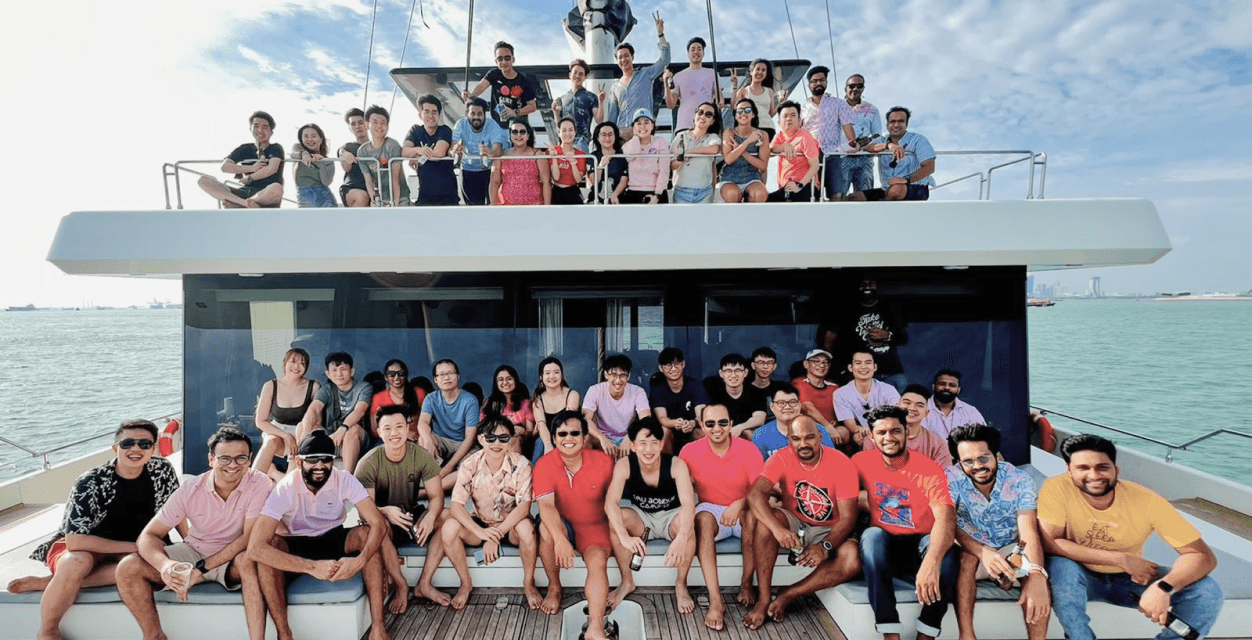 SimplifyNext team members on a boat, enjoying a daytime cruise. The boat has two levels, with team members on the upper and lower decks facing the camera for the photo. In the distance, the skyline of Singapore is visible.