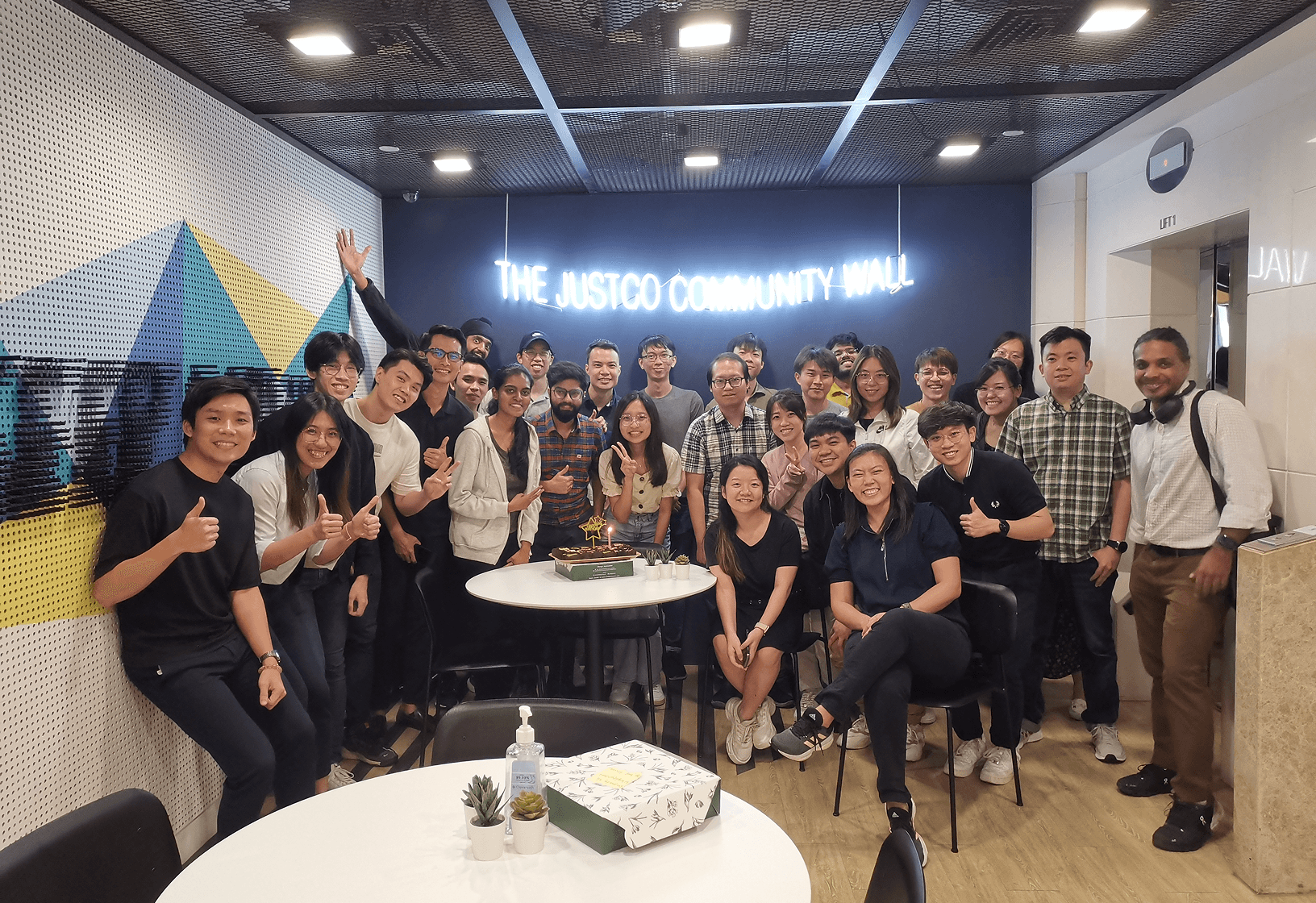 SimplifyNext team members standing together, celebrating a birthday. The team members are smiling at the camera several of them have their thumbs up.  A cake with a candle is lit in front of the team members, Behind them is a neon sign that says 'The Justco Community Hall'.