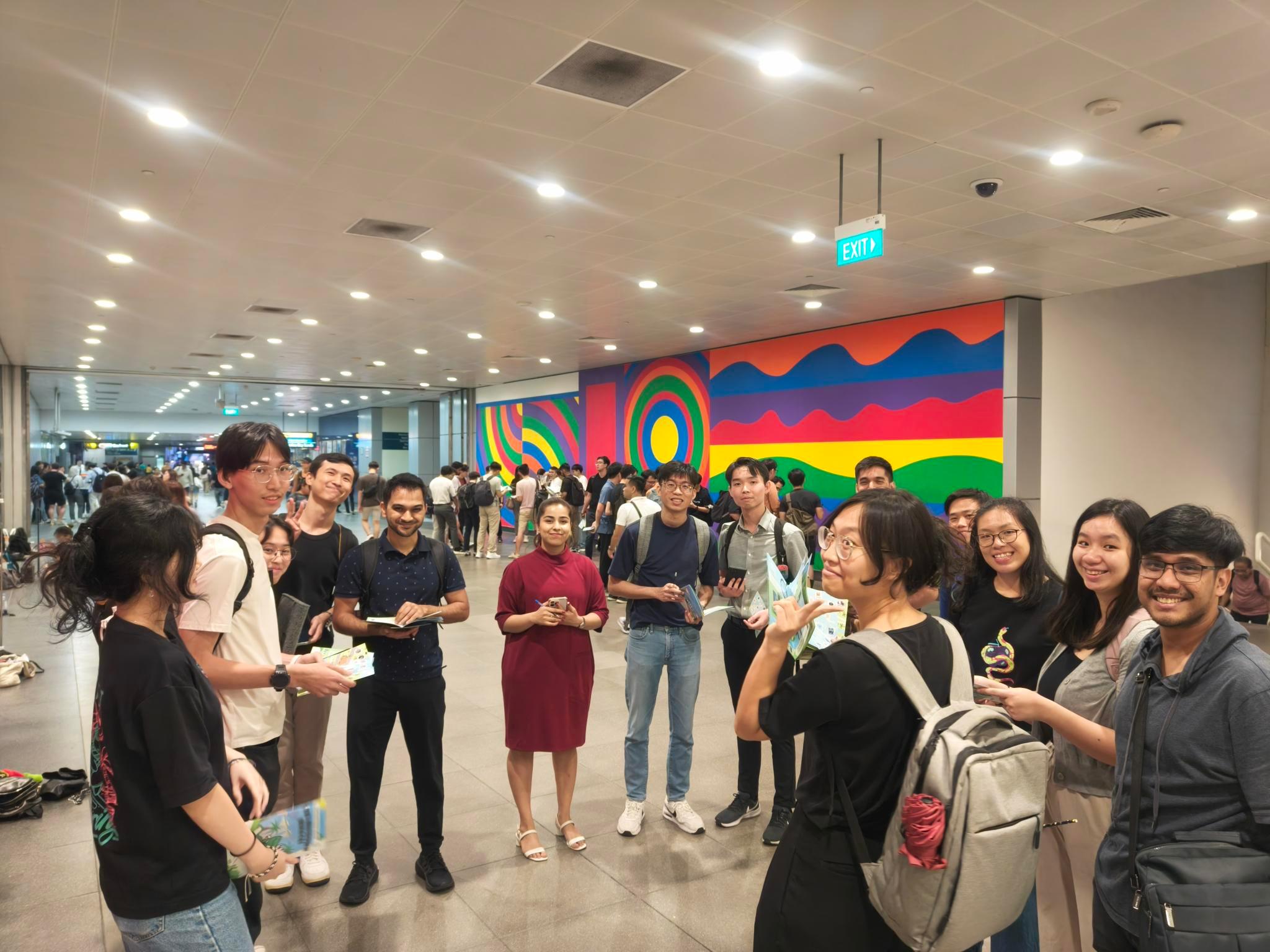 SimplifyNext team members standing in a circle, listening to a facilitator in a team building activity. In the background, several other teams are visible, engaged in the same activity with their respective facilitator. A large wall art is visible in the background. The photo is taken at Harborfront Station, Singapore.